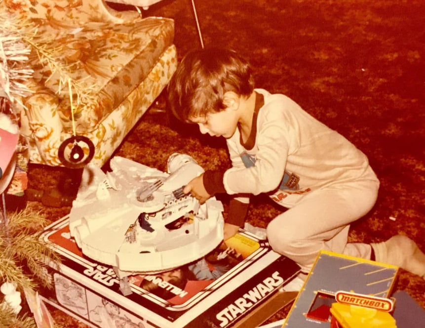 A young child in pajamas excitedly plays with a Millennium Falcon Star Wars toy spaceship next to a Christmas tree, surrounded by gift boxes and vintage furniture.