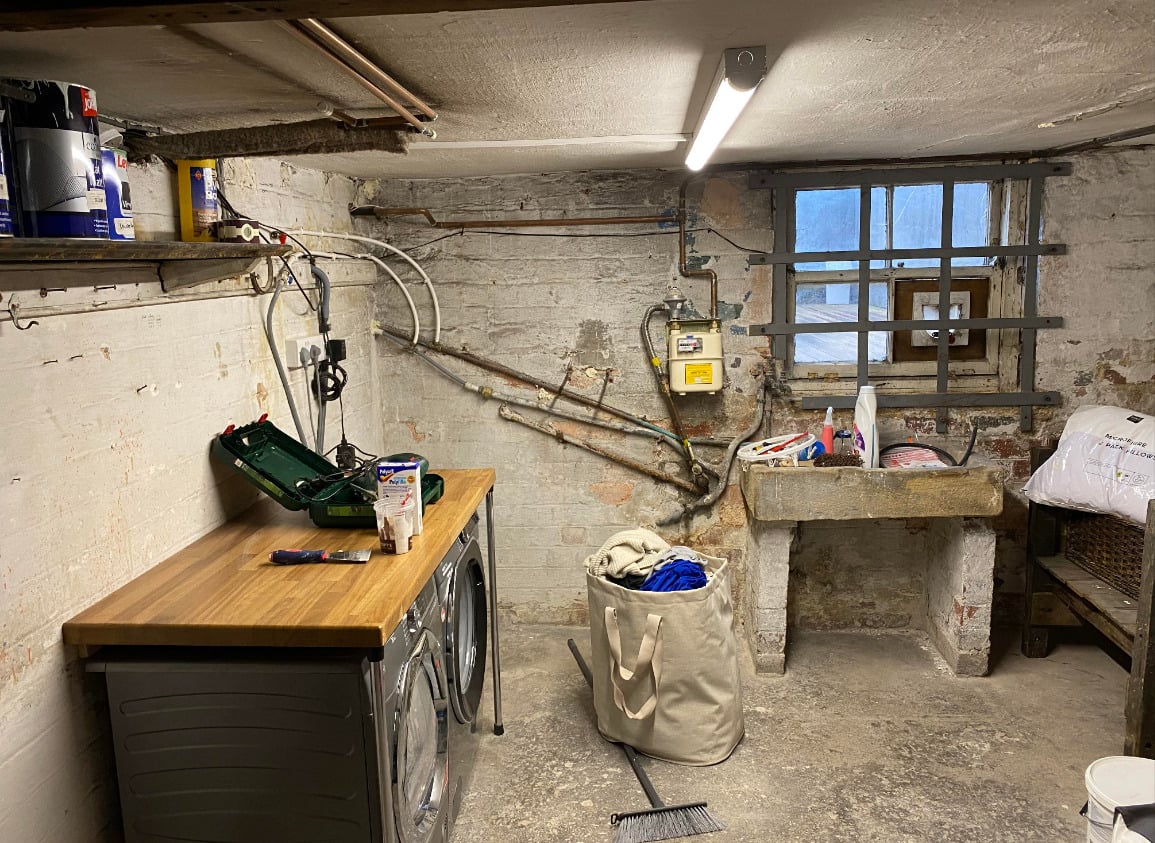 A basement laundry room with exposed pipes, a wooden counter over a washer and dryer, a sink, cleaning supplies, a laundry basket, broom, and a barred window letting in natural light.