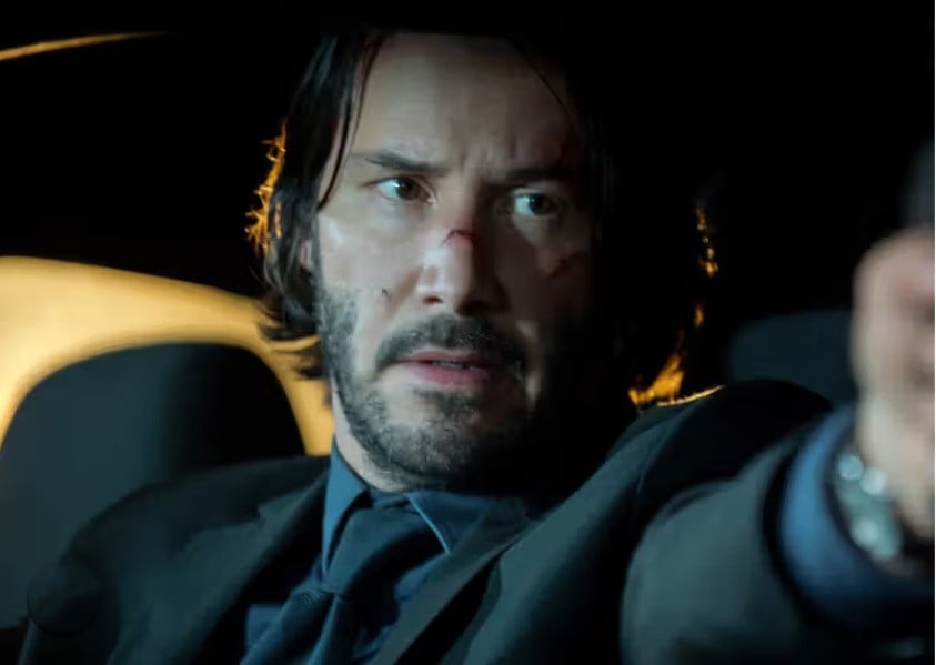 A man with scruffy hair and a beard, wearing a suit and tie, sits in a car at night with a serious expression, pointing something out of view. His face has minor cuts and bruises.