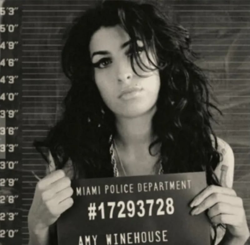 A woman with long, dark, curly hair holds a Miami Police Department sign with the number 17293728 and the name "Amy Winehouse" in front of a height chart, mimicking a mugshot. The image is in black and white.