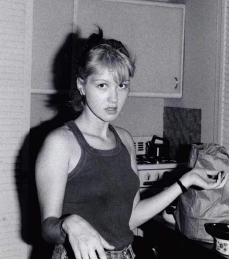 A young woman in a sleeveless top stands in a kitchen, looking at the camera with a puzzled expression. She has one hand raised and appears mid-gesture. There are cabinets, a stove, and a paper bag in the background.