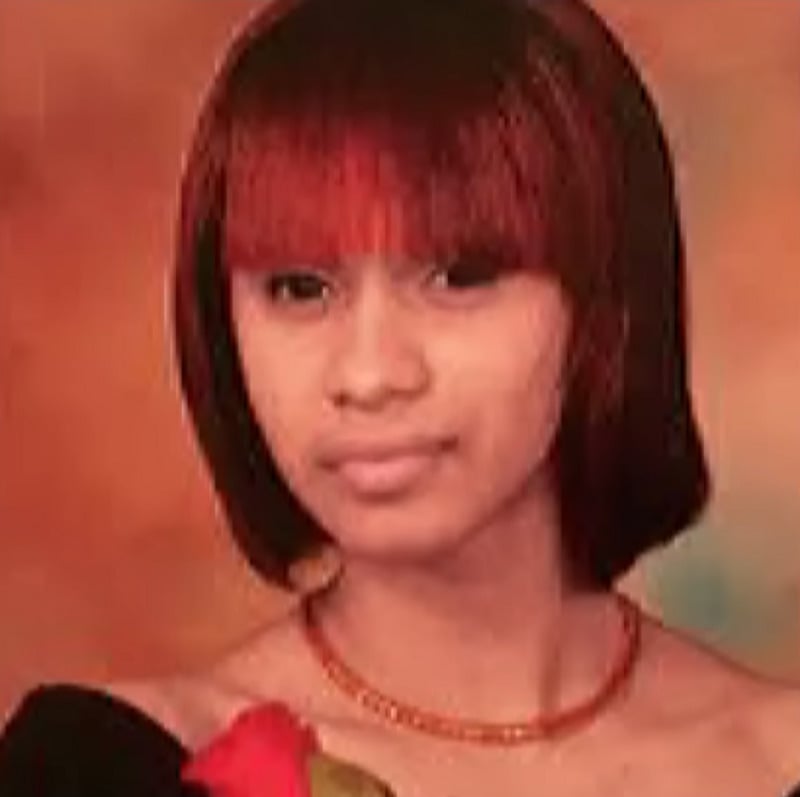 A young woman with straight, shoulder-length hair with red highlights poses for a formal portrait against a warm brown background, wearing an off-the-shoulder black top and a red necklace, and holding a red rose.