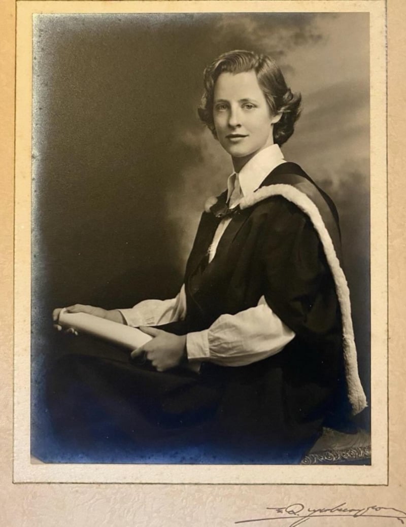 A black-and-white portrait of a young woman in academic robes holding a diploma, seated and looking at the camera with a calm expression against a cloudy studio backdrop.