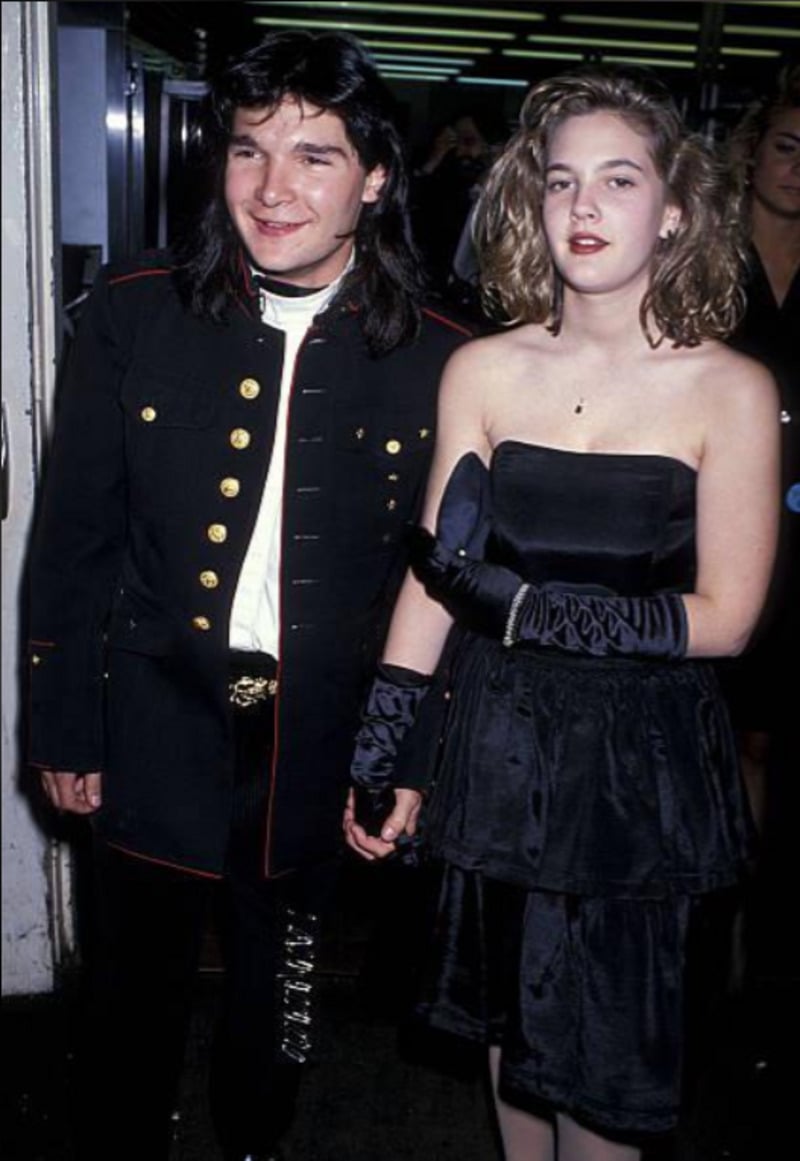A young man in a black military-style jacket and a young woman in a strapless black dress and gloves stand hand-in-hand, posing and smiling at an indoor event with people in the background.