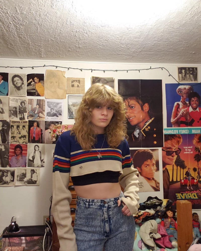 A person with wavy blonde hair wearing a striped crop sweater and high-waisted jeans stands in a bedroom decorated with numerous Michael Jackson posters and black-and-white photos on the wall.