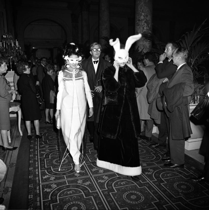 Two women in elaborate masks and costumes, one with a rabbit mask and ears, walk on a patterned carpet at a crowded, formal indoor event with elegantly dressed guests observing them. The setting appears stylish and festive.