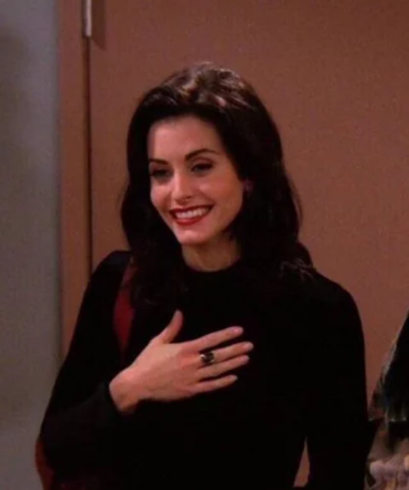 A woman with shoulder-length dark hair, wearing a black top and a ring, smiles warmly with her hand placed on her chest in a brightly lit indoor setting.
