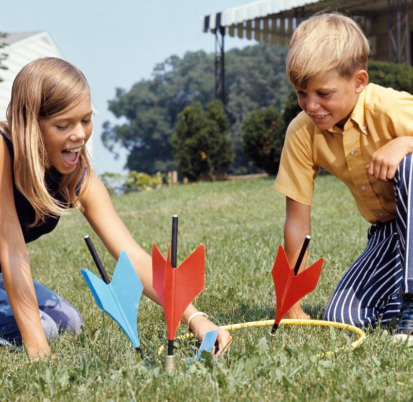 Two children kneel on grass, playing with large, colorful lawn darts and a yellow plastic ring. The girl is smiling and reaching toward a blue dart, while the boy watches and smiles. Trees and a building are visible in the background.