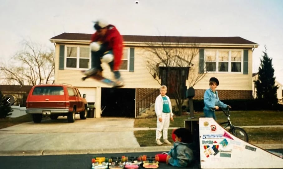 A child in a red jacket is mid-air on a skateboard above a ramp in a suburban driveway. Two kids stand nearby, one with a bike, while another sits on the ground playing with toys. A red SUV is parked by a house in the background.