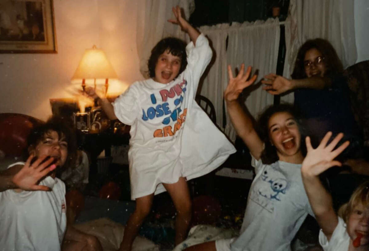 Four children are joyfully posing in a living room at night, with one child standing on a chair and others waving their hands toward the camera. The room is warmly lit by a lamp, and there are curtains and scattered belongings.