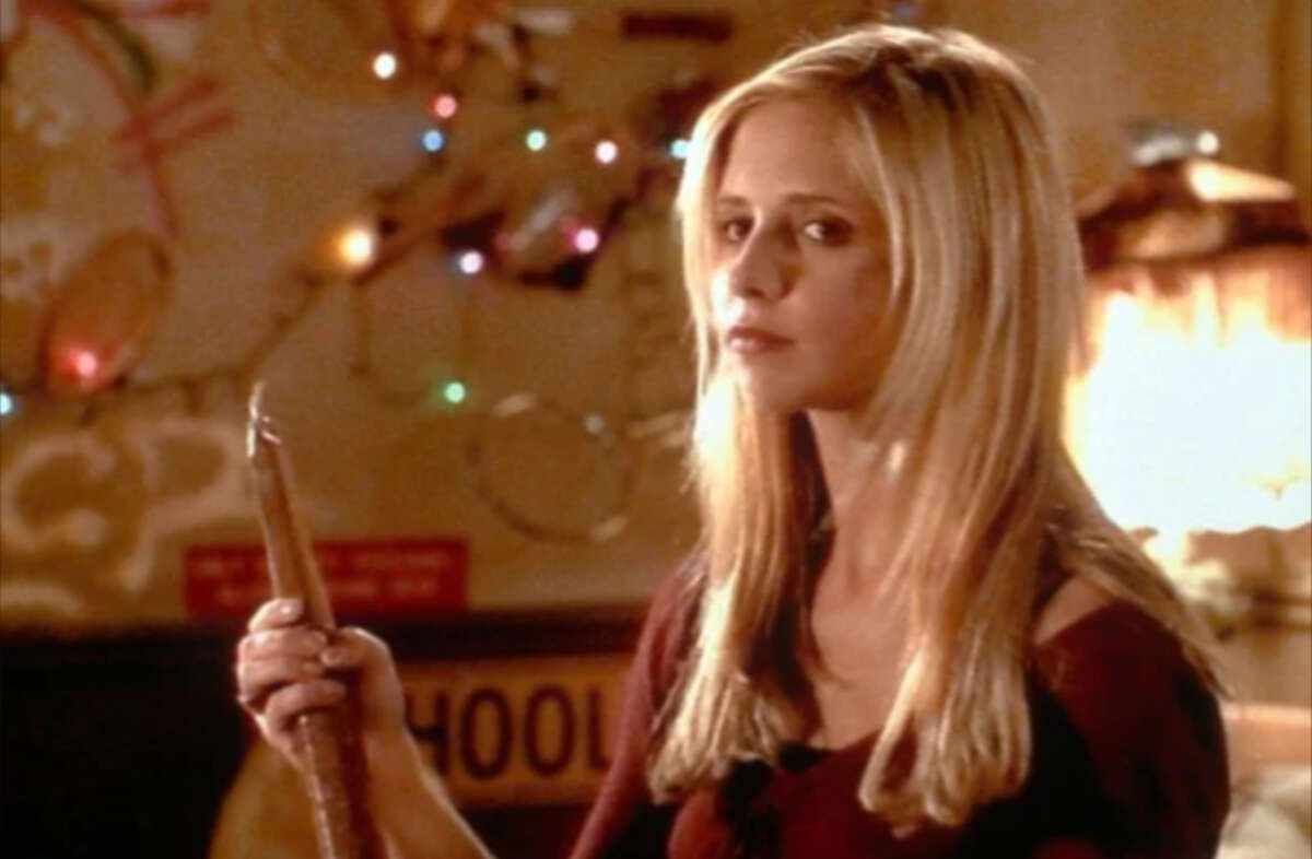 A young woman with long blonde hair holds a wooden stake and looks serious. She stands indoors with colorful string lights and a guitar hanging on the wall behind her.