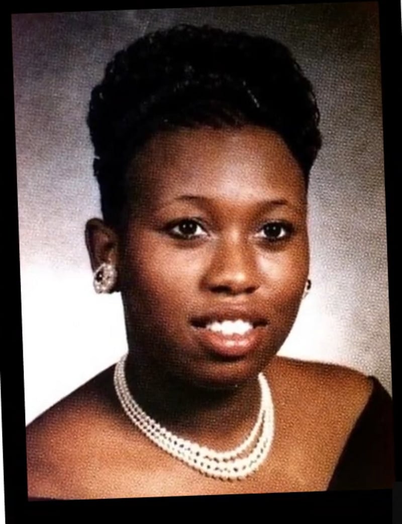 A young woman poses for a formal portrait, wearing a dark off-the-shoulder top, pearl necklace, and matching earrings. She has short, styled hair and is smiling softly against a neutral background.