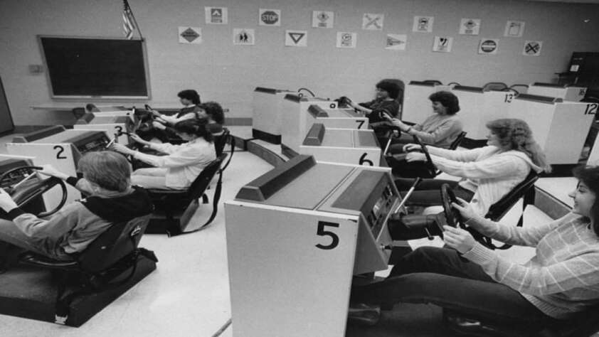 Black and white photo of students in a classroom, each seated at a driving simulator station with steering wheels, practicing driving skills. Road signs are displayed on the back wall.