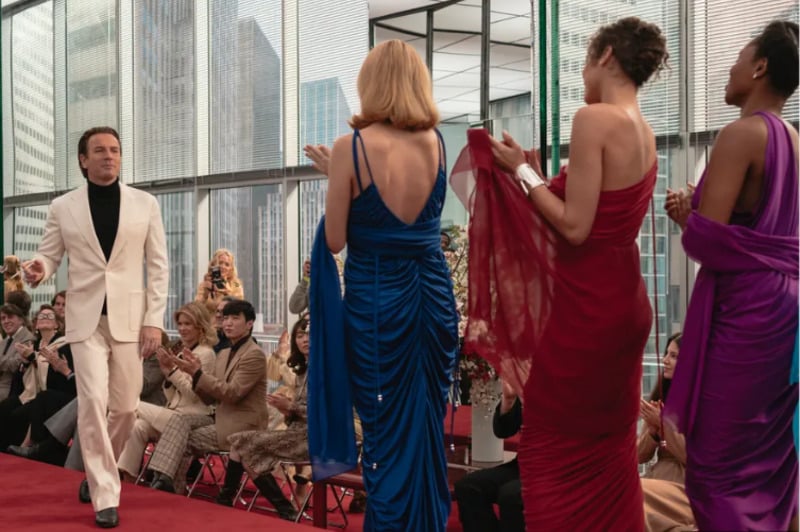 A man in a white suit walks on a red carpet runway toward three women in elegant gowns, as an audience seated on both sides applauds in a modern, glass-walled city building.