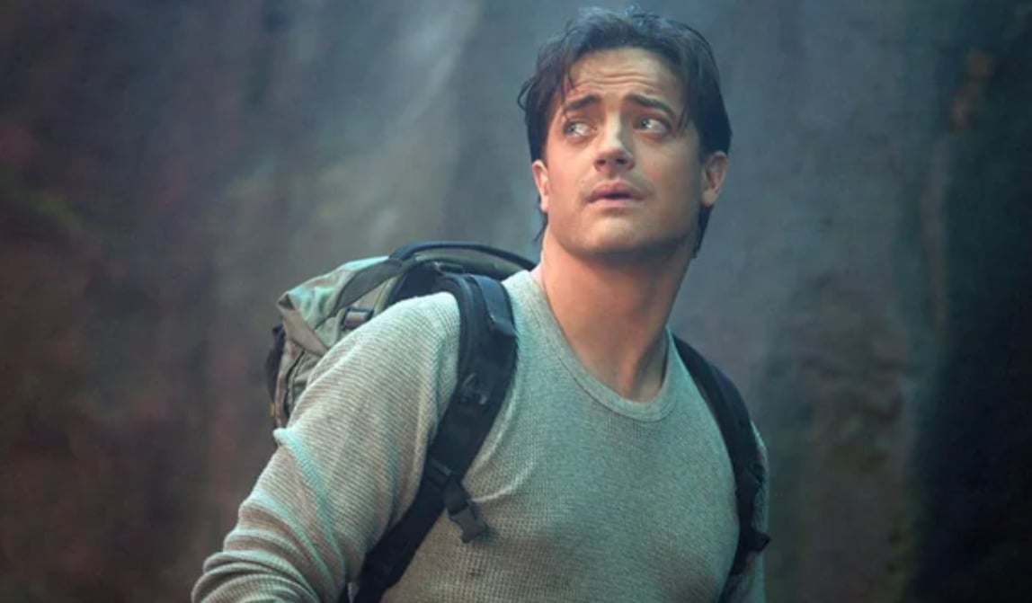A man with short dark hair, wearing a gray long-sleeve shirt and a backpack, looks up with a concerned expression. The background appears rocky and outdoors.