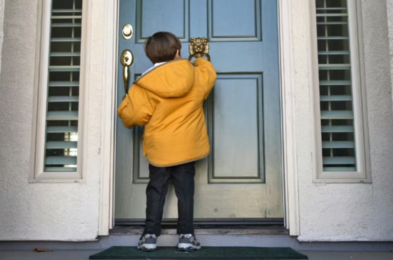 A young child in a yellow coat stands on a doorstep, seen from behind, knocking on a blue front door of a house with white walls and shuttered windows.