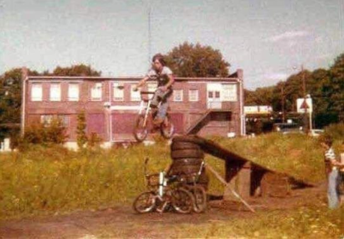 A young person on a bicycle jumps off a wooden ramp over a stack of tires and other bikes, with an old brick building and trees in the background. Another person stands nearby watching.