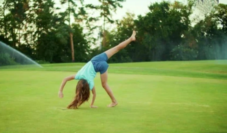 A girl with long hair, wearing a blue shirt and shorts, performs a cartwheel barefoot on a grassy field with sprinklers and trees in the background.