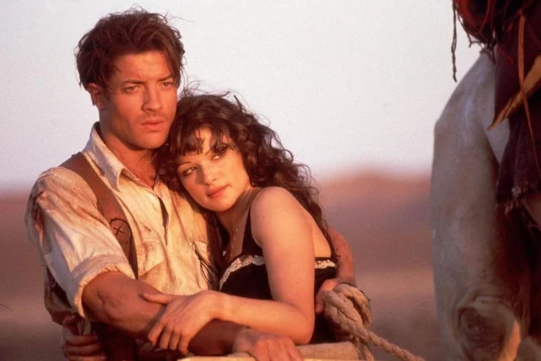 A man and a woman stand close together in an embrace at sunset. The man looks off into the distance while the woman rests her head on his chest, gazing forward. Both appear dusty and are dressed in old-fashioned, adventurer-style clothes.