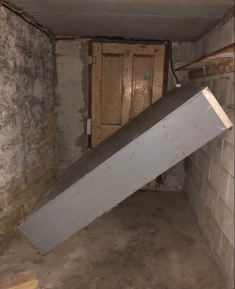 A large, gray rectangular box leans diagonally against a rough, cinder block wall in a dim, unfinished basement with a weathered wooden door in the background.