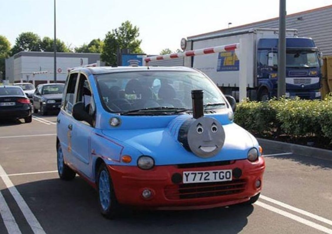 A small car decorated to look like Thomas the Tank Engine, with a blue body, red accents, and a large smiling face and chimney on the hood, parked in a lot near trucks and other vehicles.