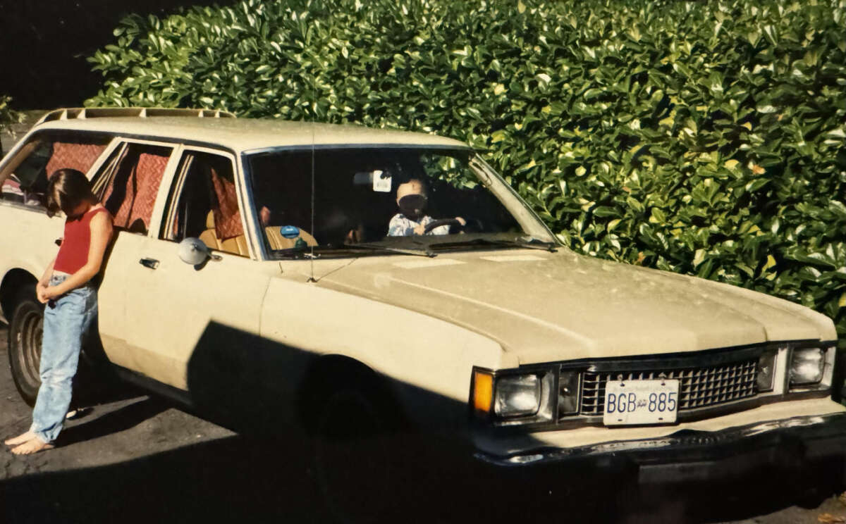 A girl in a red top and jeans stands barefoot next to an old beige station wagon parked by a tall green hedge. Two children are visible inside the car, one behind the wheel.