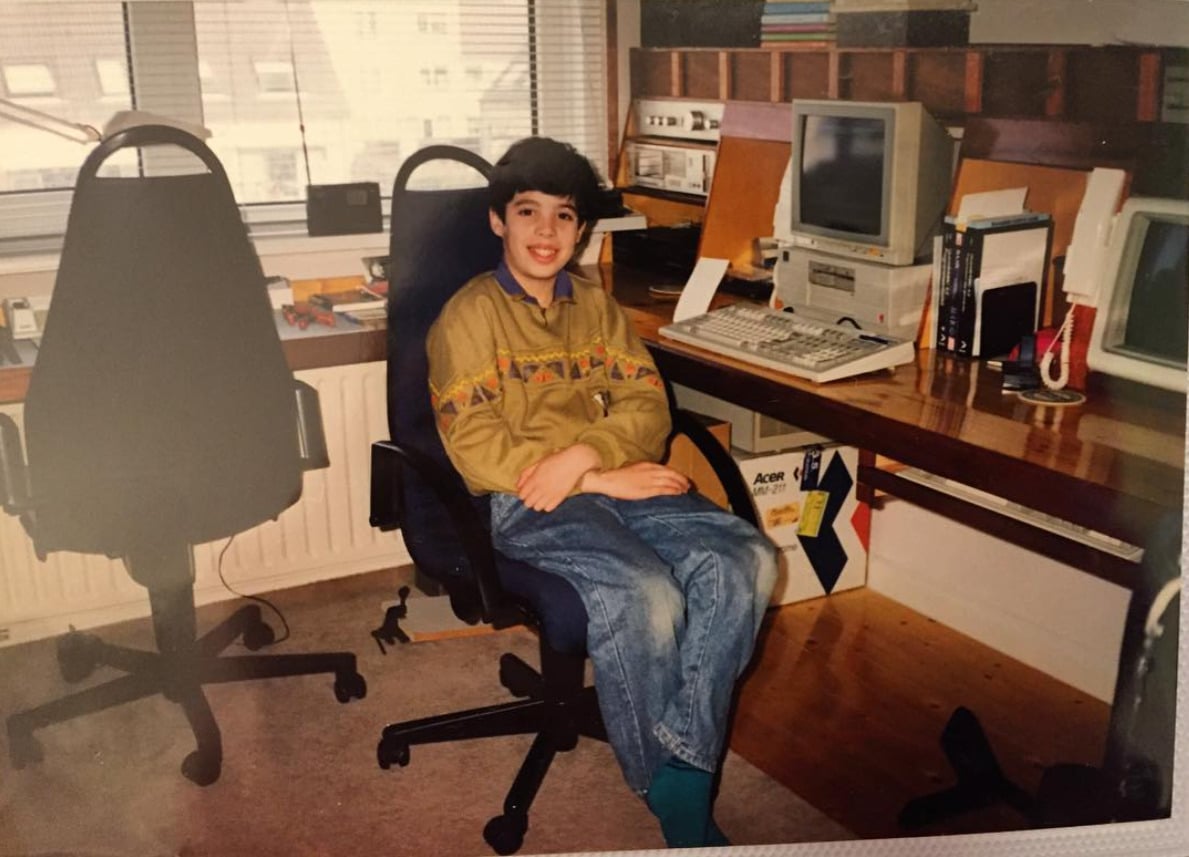 A smiling boy sits on an office chair in a vintage home office with a 1990s computer on a wooden desk, books, and shelves in the background. A window lets in natural light. Another empty chair is nearby.
