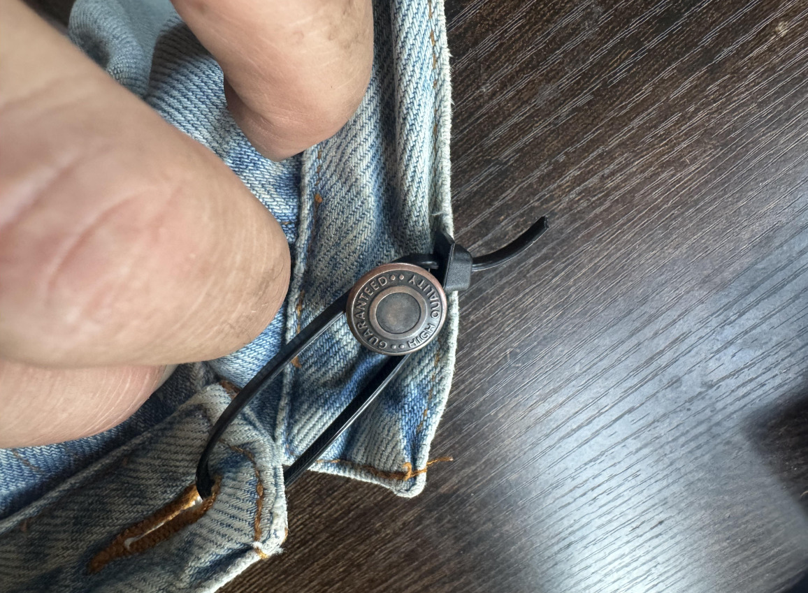 A close-up of a hand holding the waistband of blue denim jeans, secured with a black zip tie looped through the buttonhole and around the metal button, on a dark wooden surface.