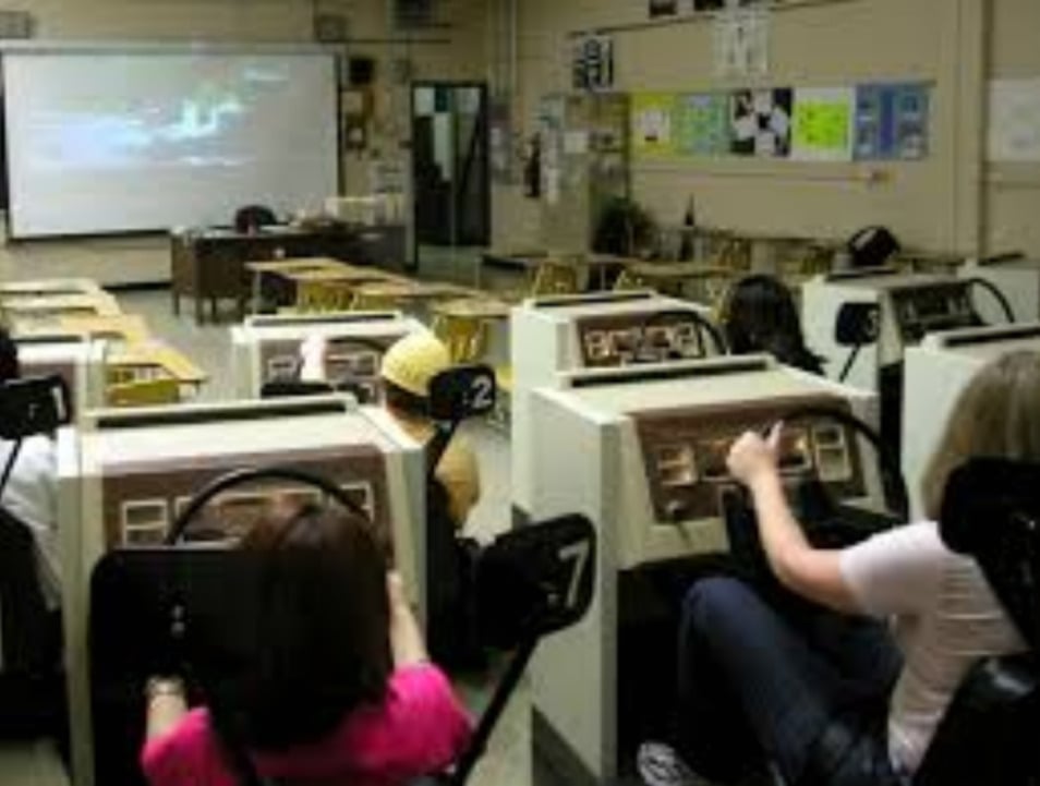 Several people sit in individual driving simulators facing a large screen at the front of a classroom, each holding a steering wheel as part of a driver’s education lesson.