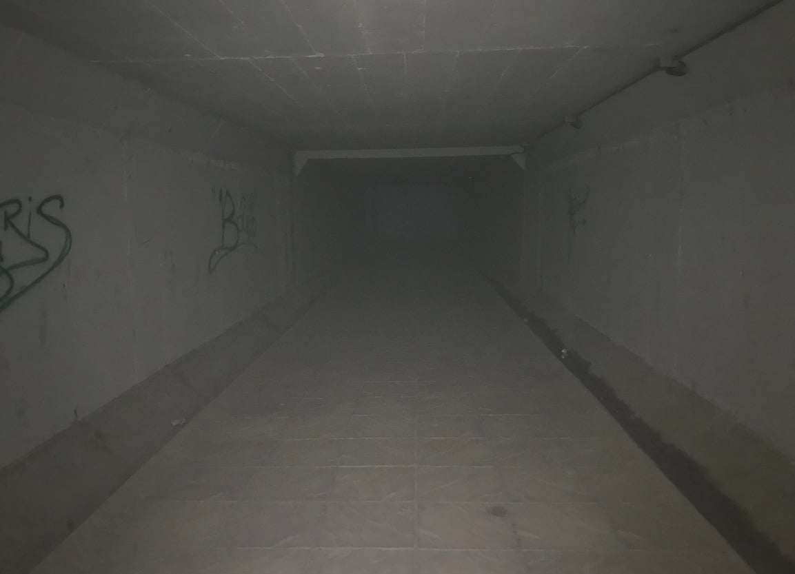 A dark, empty tunnel with concrete walls covered in graffiti and a tiled floor, fading into darkness in the distance. Dim lighting creates an eerie, unsettling atmosphere.