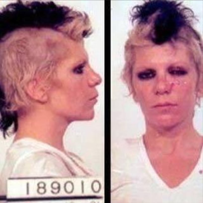 A person with short, messy, bleach-blond and black hair is shown in side and front mugshot photos, wearing a white shirt and dark eye makeup, with the number 189010 visible on the side image.