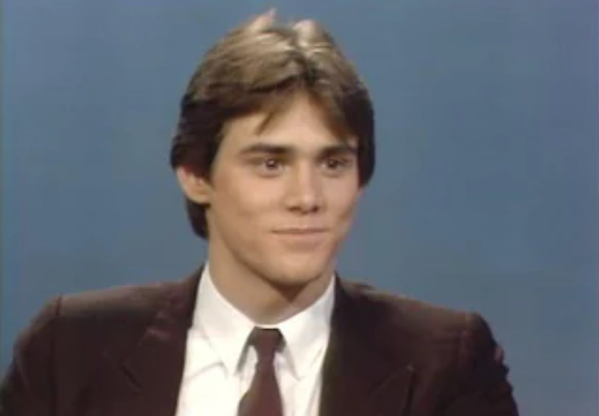 A young man with short brown hair wearing a dark suit, white shirt, and dark tie sits in front of a plain blue background, looking slightly to the side and smiling softly.