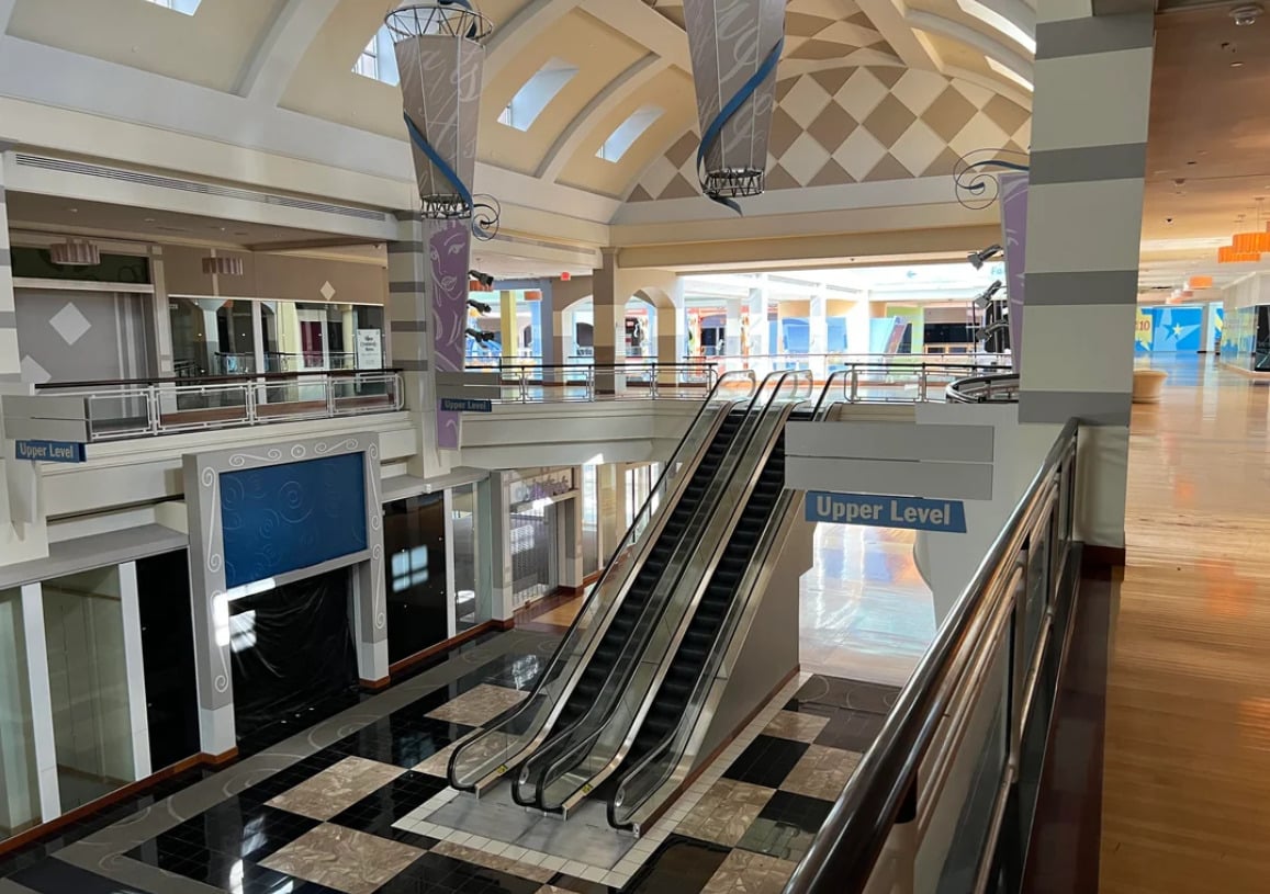 An empty, well-lit shopping mall interior with closed storefronts, escalators leading to an upper level, and decorative banners hanging from a high arched ceiling. No people are visible.