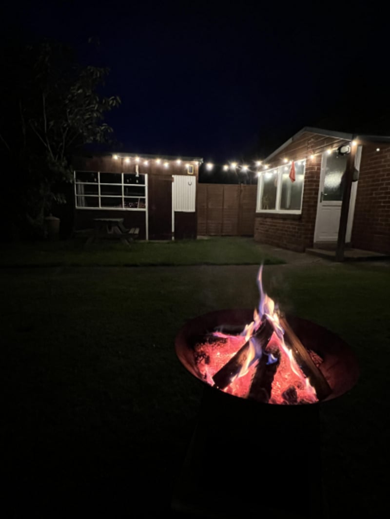 A fire pit with glowing logs burns in the foreground of a dark backyard. In the background, string lights illuminate a small building with a picnic table and a brick house with lit windows and doors.