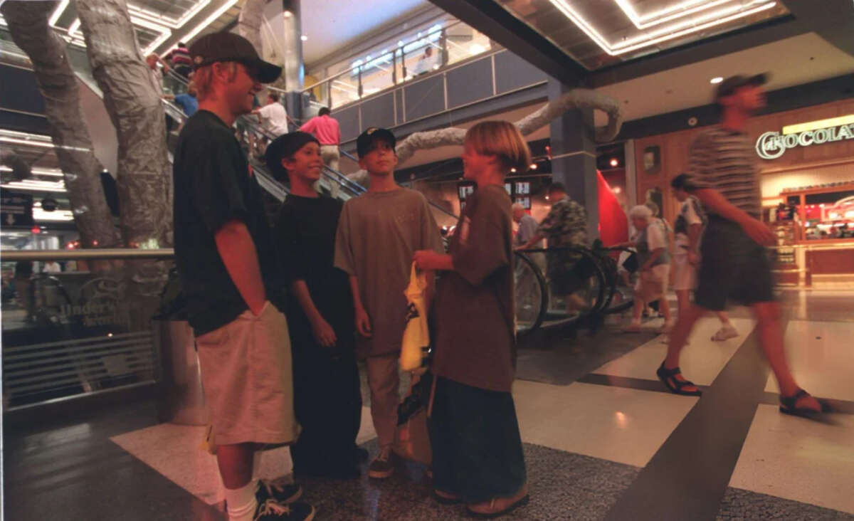 Four kids in baggy clothes and hats stand talking inside a shopping mall, with an escalator and store signs in the background. Other shoppers walk by, and the scene is lit with bright overhead lights.