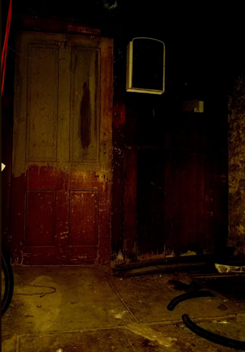 A dimly lit, old wooden door stands in a worn, grimy room with a dirty floor, scattered debris, and dark walls. A white speaker or small box is mounted above the door. The atmosphere feels eerie and neglected.