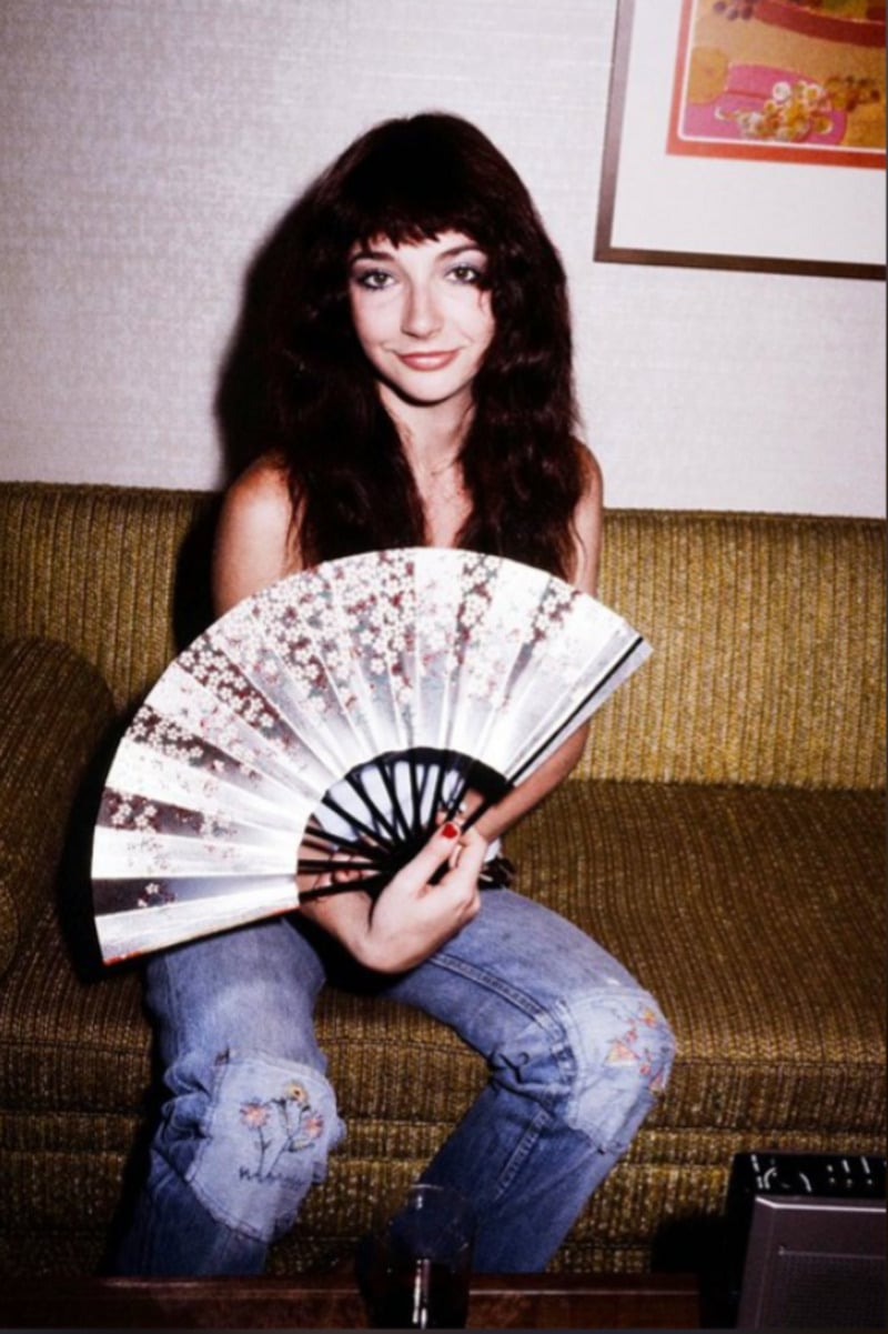 A woman with long brown hair sits on a green sofa, smiling and holding an open decorative fan. She wears a white top and blue jeans with embroidered flowers, and there is a framed artwork on the wall behind her.
