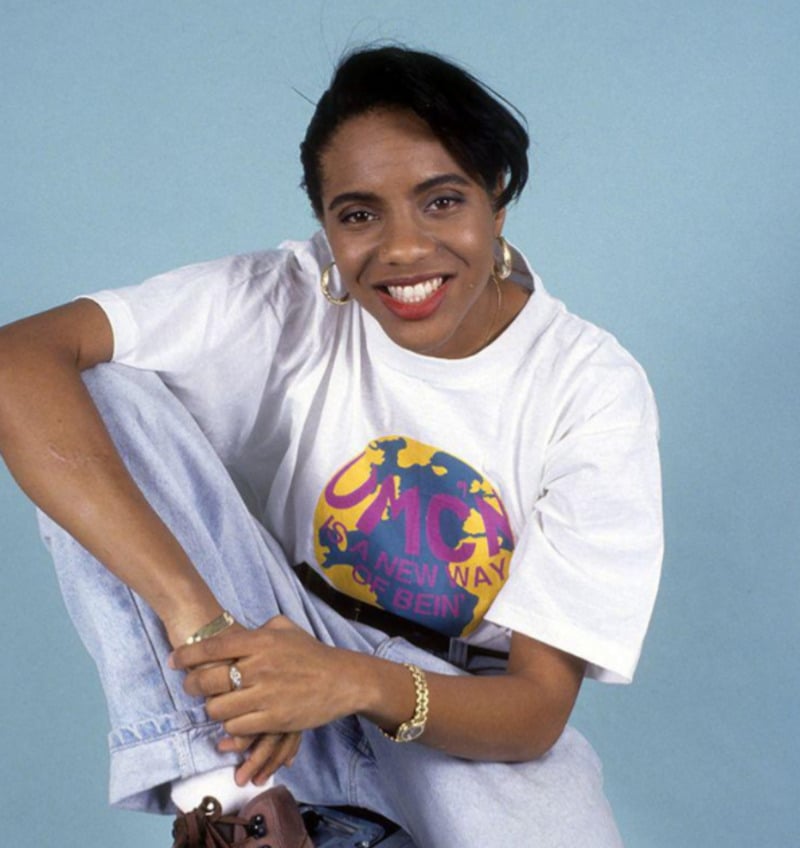 A woman with short black hair smiles at the camera, wearing hoop earrings, a white t-shirt with colorful graphics, light jeans, and sneakers, posing against a light blue background.