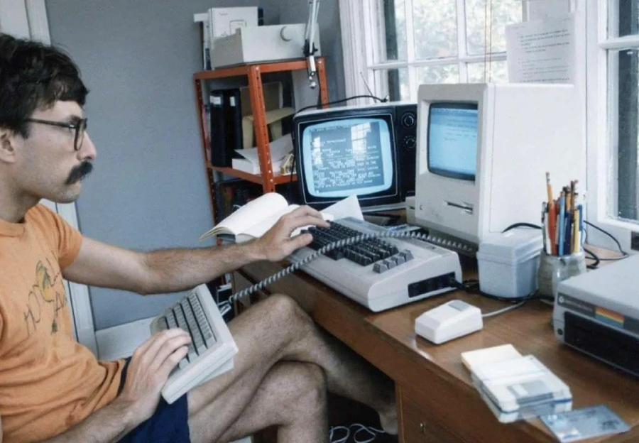 A man with glasses and a mustache works at a desk with vintage computers, floppy disks, and two monitors. He is typing on one keyboard while holding another, surrounded by office supplies and papers.