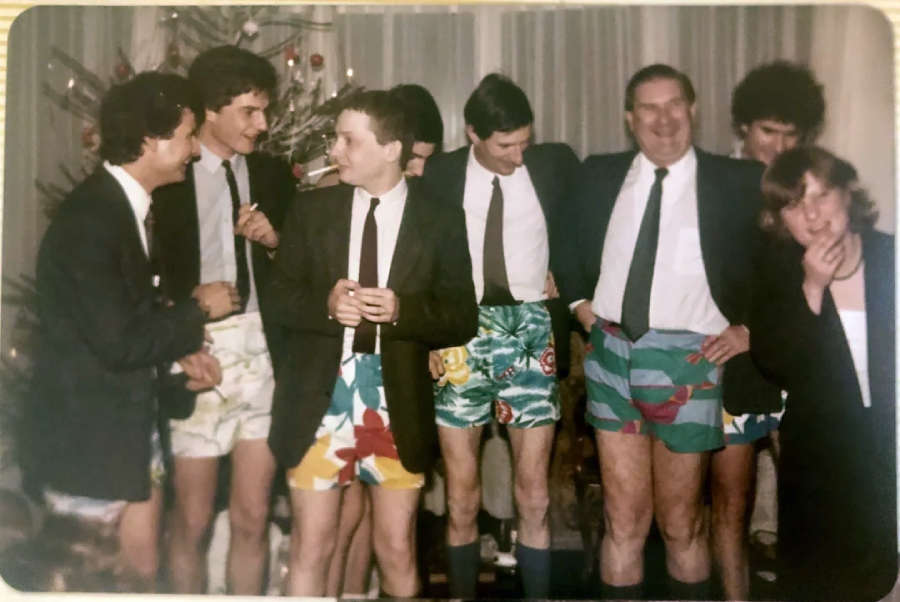 A group of six men in suits and colorful shorts and one woman stand together indoors, smiling and laughing, with a decorated Christmas tree in the background.