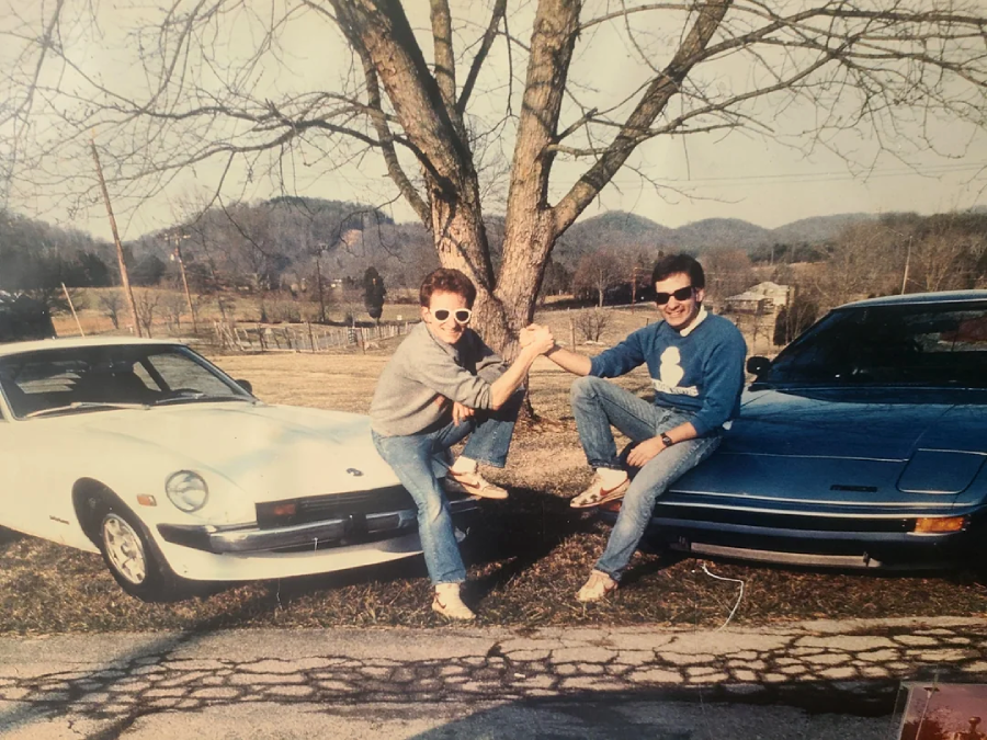 Two people in sunglasses pose playfully, clasping hands between a white classic car and a blue sports car, with a leafless tree and rolling hills in the background on a sunny day.