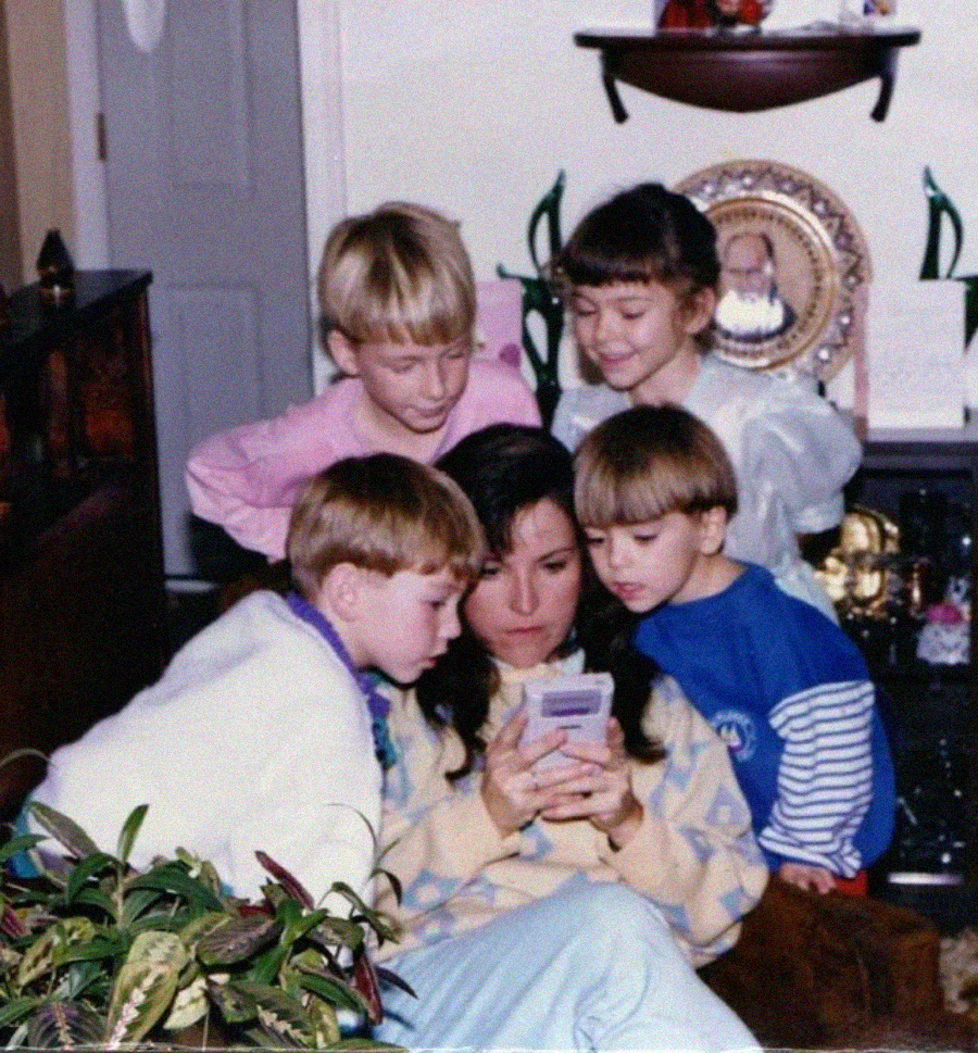 A woman sits on a couch holding a handheld game console while four young children crowd around her, eagerly looking at the screen. They are indoors, and there are houseplants and decorations in the background.