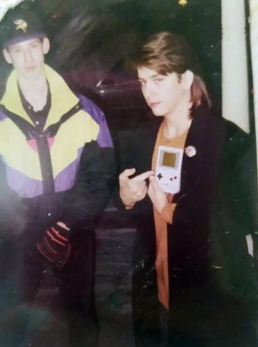 Two young men stand together; one wears a colorful jacket and cap, the other holds a classic Game Boy, pointing at it and looking at the camera. The photo has a vintage, 1990s feel.