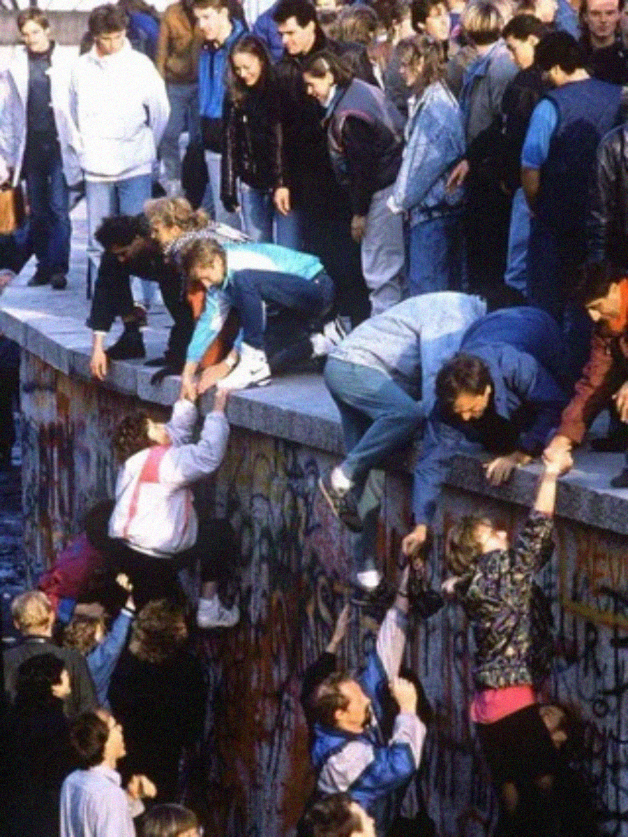 A crowd of people helps each other climb over a graffiti-covered concrete wall, with some already on top and others reaching up from below.