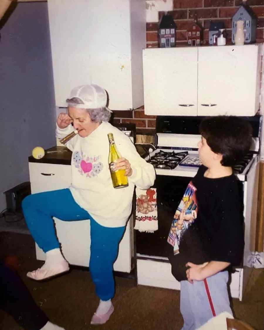 An elderly woman in a white cap, sweatshirt, and blue pants dances playfully in a kitchen while holding a drink, as a young child in a graphic t-shirt watches and smiles.
