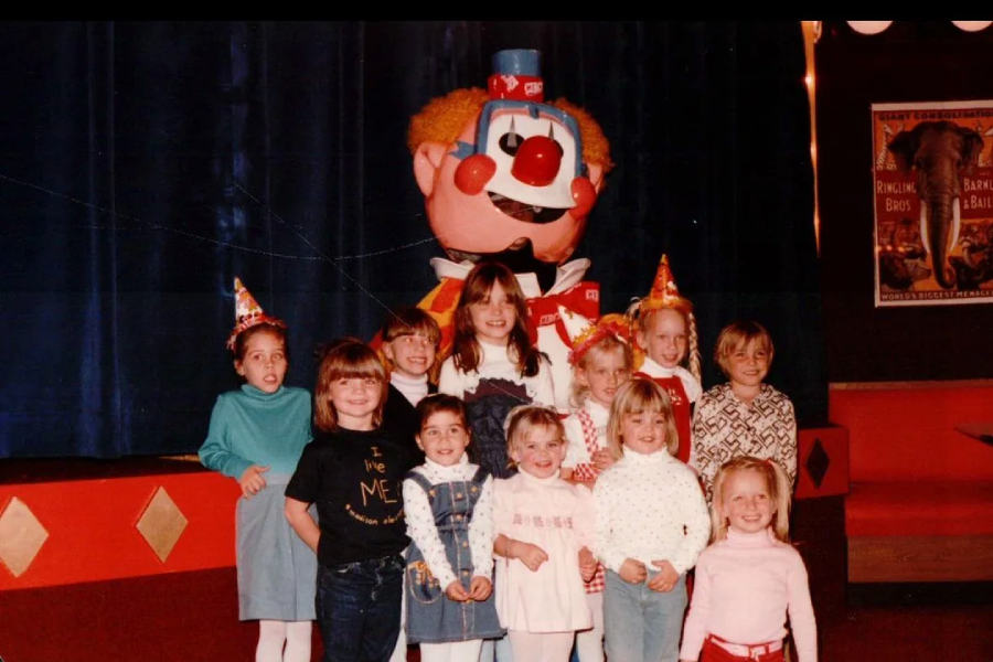 A group of children wearing party hats pose and smile in front of a large clown figure on a stage. The background is decorated with red and blue, and posters are visible on the wall to the right.