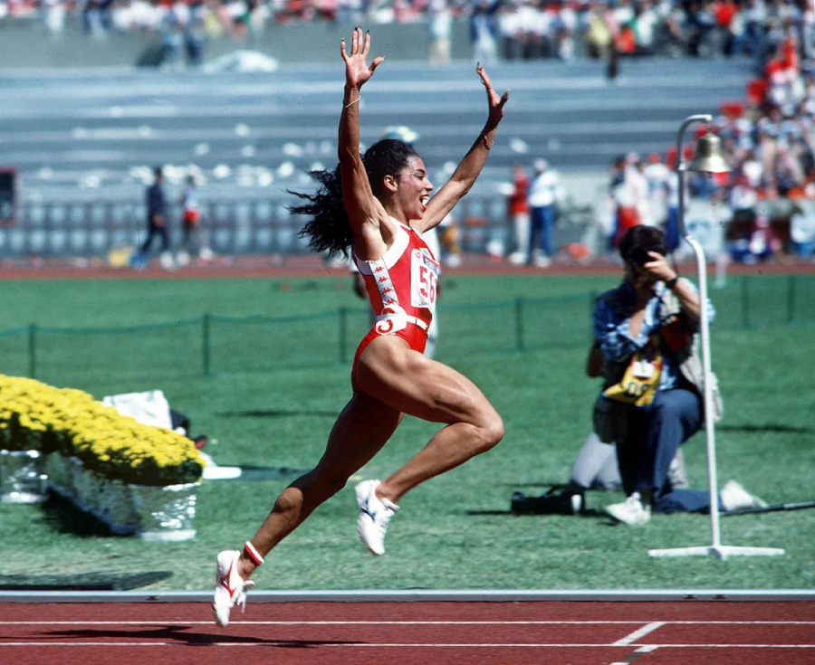 A female track athlete in a red and white uniform sprints across the finish line with arms raised in victory on an outdoor track, as spectators and photographers watch in the background.