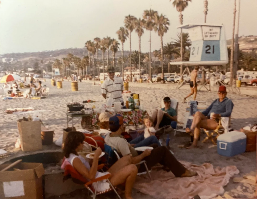 A group of people sits on beach chairs around a table with food and drinks on a sandy beach. Palm trees, a lifeguard tower, and other beachgoers are visible in the background.