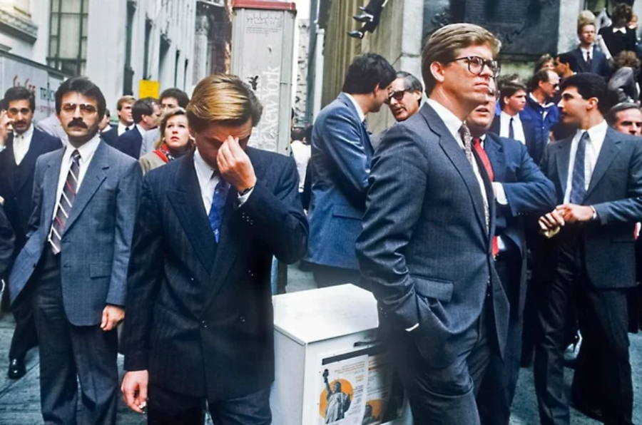A busy city street scene shows several men in business suits walking or standing, some appearing stressed or deep in thought, in an urban area lined with tall buildings.