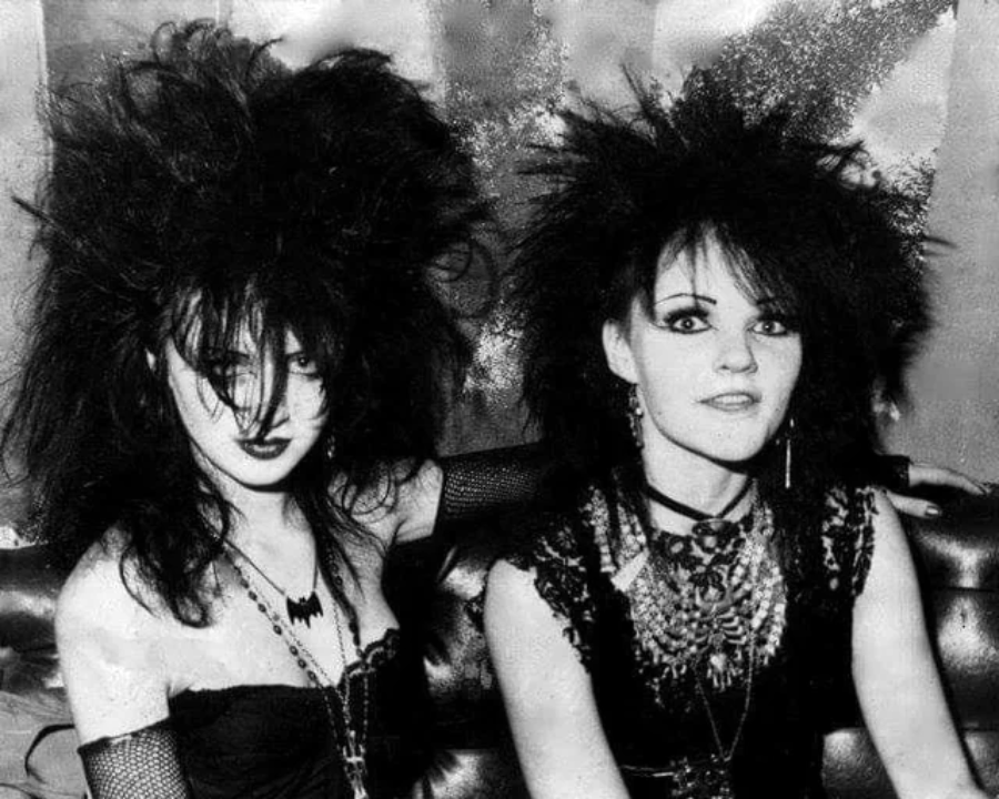 Two women with dramatic, teased black hair and heavy makeup sit on a couch. They wear black gothic clothing, fishnet gloves, bold jewelry, and dark lipstick, embodying a punk or goth style.
